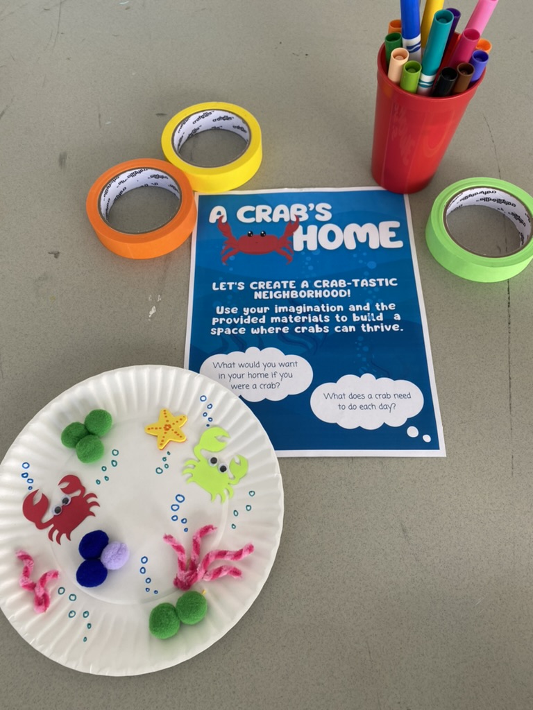 A table with crafting supplies, and a worksheet titled "A Crab's Home." A paper plate decorated with crabs and pipe cleaners sits next to it.