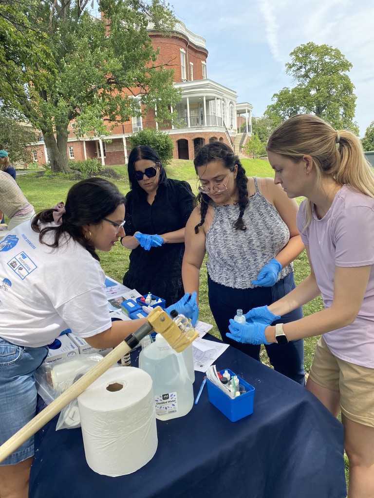 Three teachers are wearing blue latex gloves, one holding a small bottle. A scientist also wearing gloves talks to them.