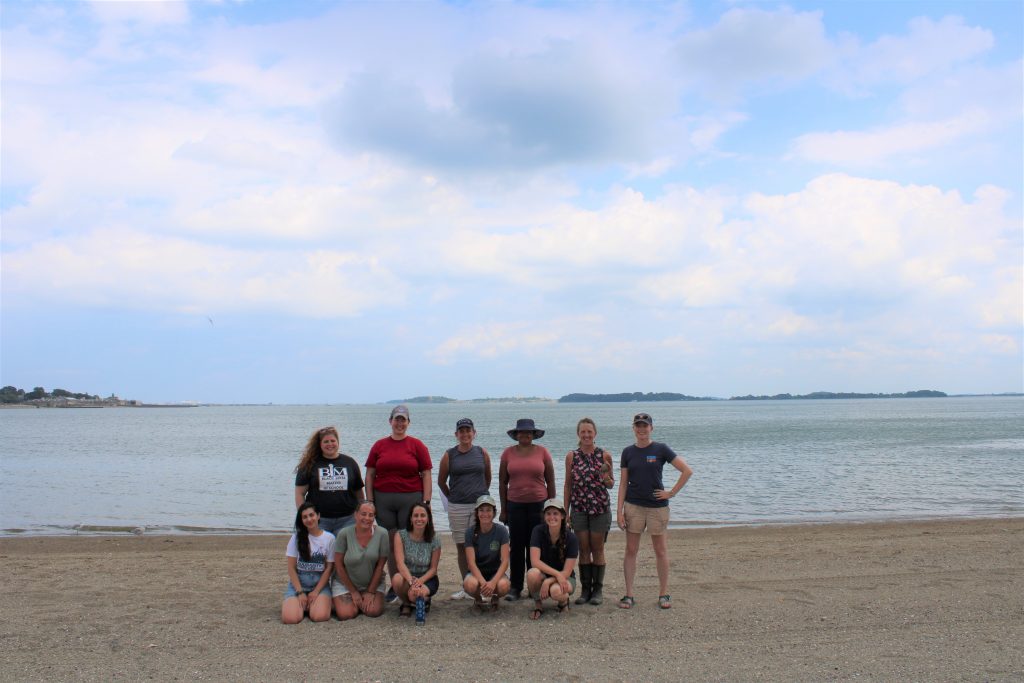 A group of adults standing together on a beach, smiling at the camera.