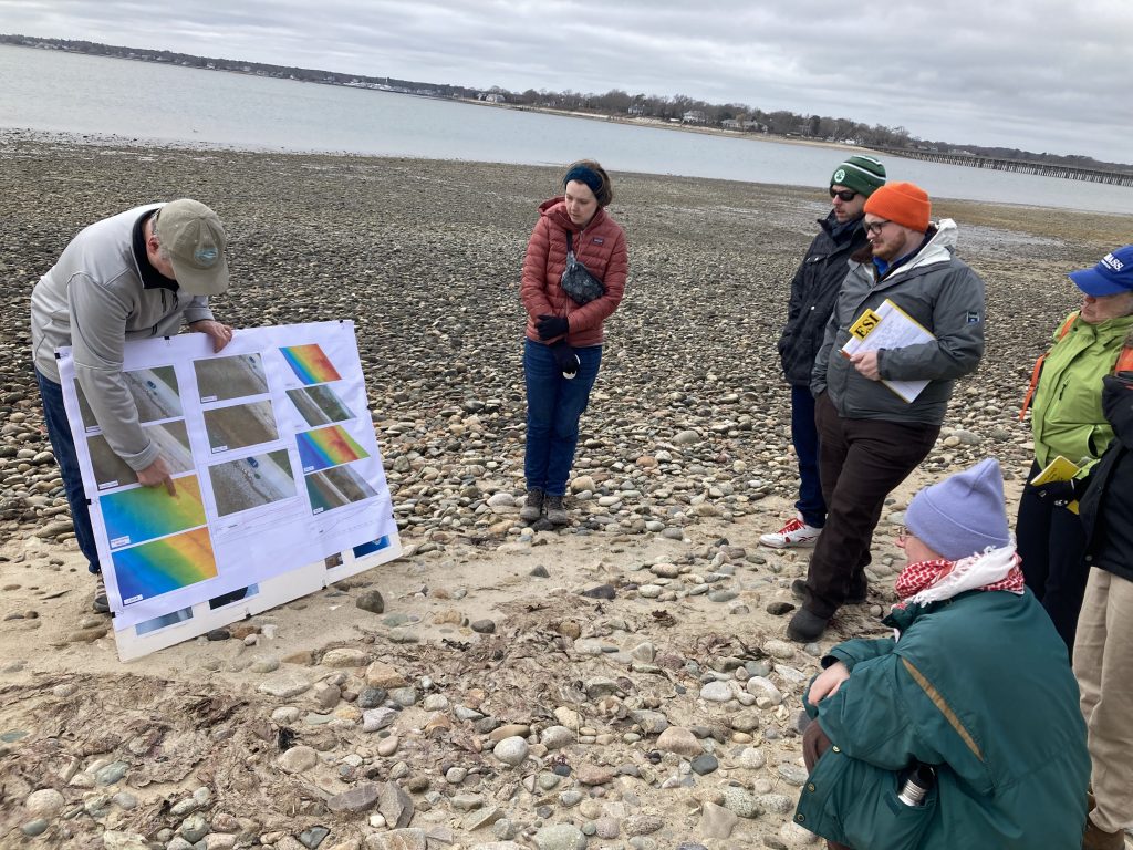 A group of people gathered on a beach, while a scientist points to scientific figures on a poster.