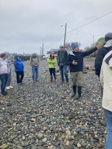 A group of people gathered on a cobble beach, while a speaker points off screen.